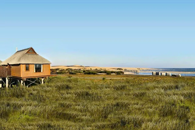 Hölzernes Pfahlhaus über Küstengras mit Dünen, Pier und Meer bei klarem Tageslicht.