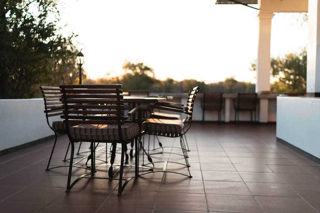 Leere Terrasse mit Metallstühlen und Tisch im warmen Abendlicht, Bäume im Hintergrund.