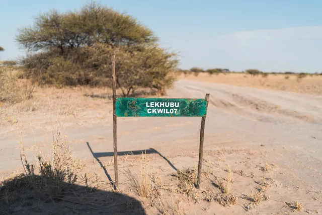 Verwittertes grünes Wegschild an einer Sandpiste in trockener Savannenlandschaft bei Sonnenschein.