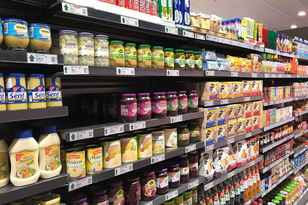 Supermarktregal mit Gläsern, Dosen und Kartons unter heller Deckenbeleuchtung.