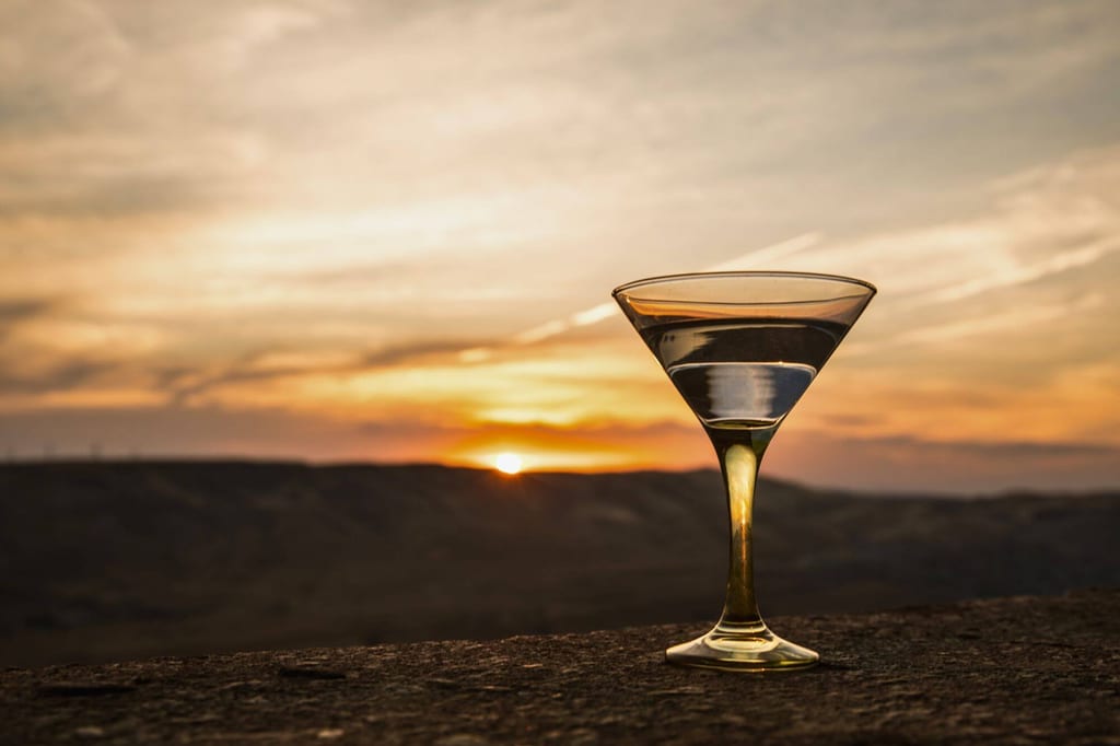 Cocktailglas mit klarem Getränk auf Felskante bei Sonnenuntergang, warme Abendstimmung