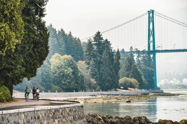 Uferpromenade mit Radfahrern, Bäumen und großer Hängebrücke bei bewölktem Tageslicht.