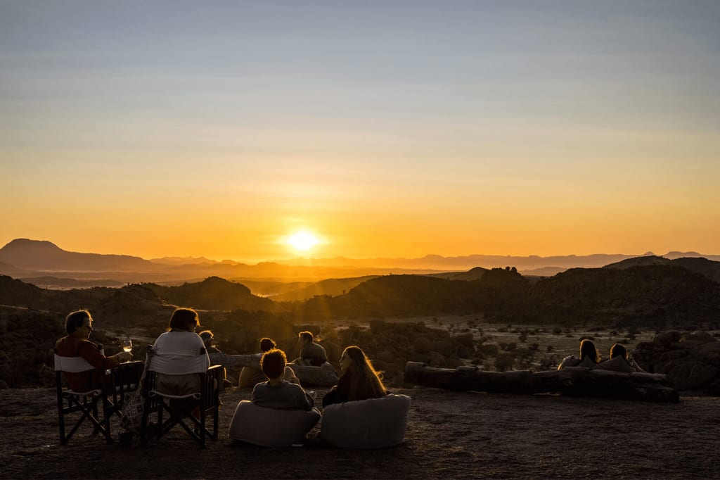 Menschen sitzen bei goldenem Sonnenuntergang auf einer Anhöhe und schauen über eine felsige Landschaft.