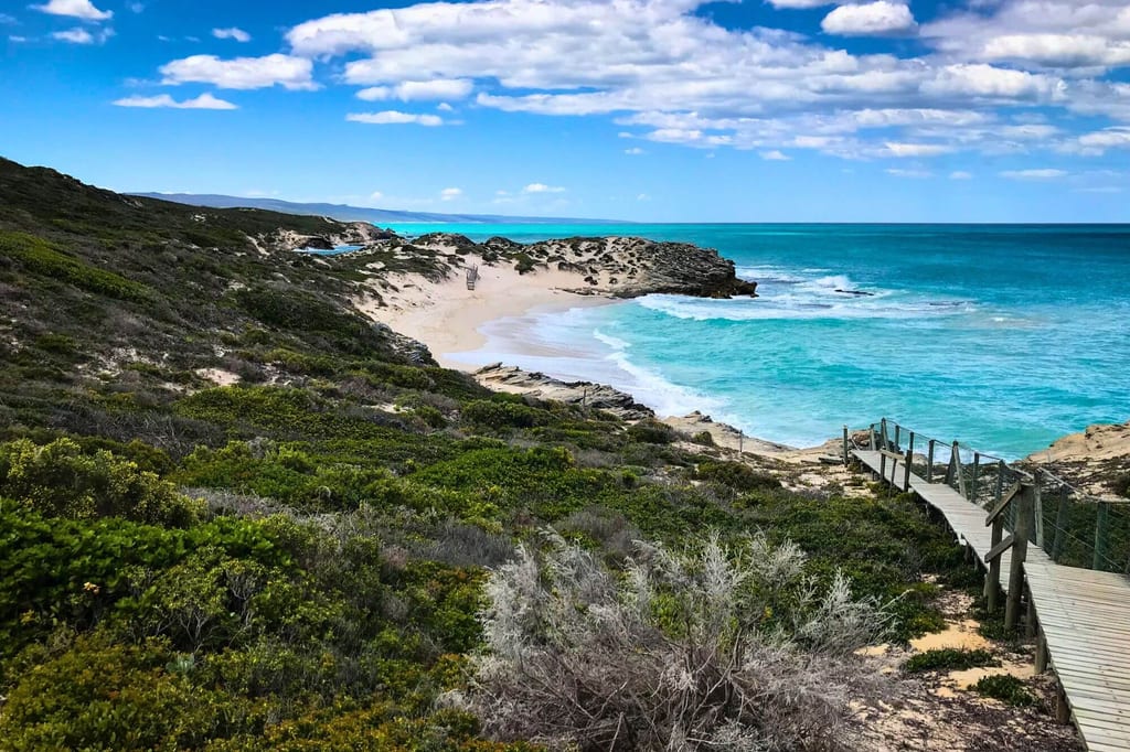 Blick über Dünen zu einem Sandstrand mit türkisfarbenem Meer und Holzsteg unter teils bewölktem Himmel.