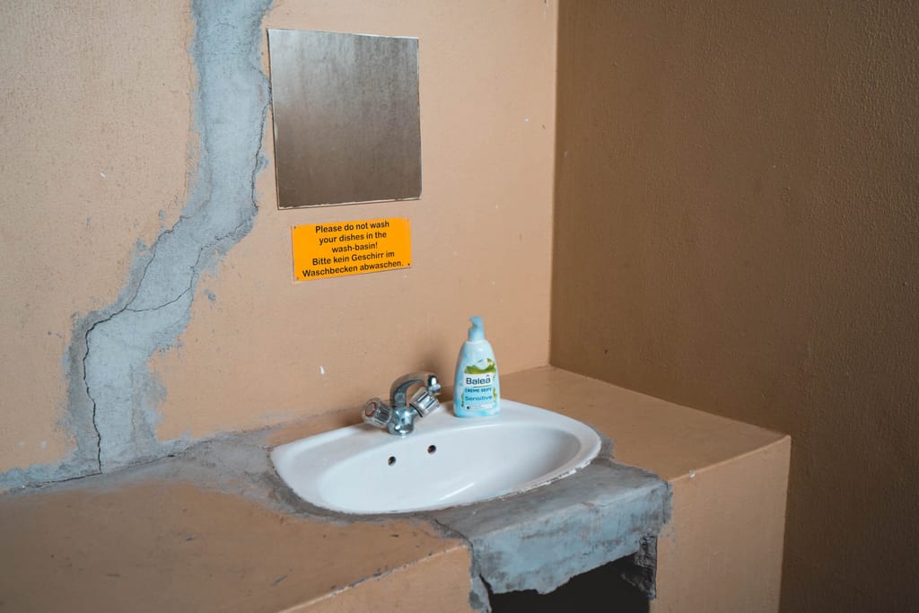 Frontalaufnahme eines einfachen Waschbeckens mit Seifenflasche, Spiegel und rissigem Putz an beige Wand.