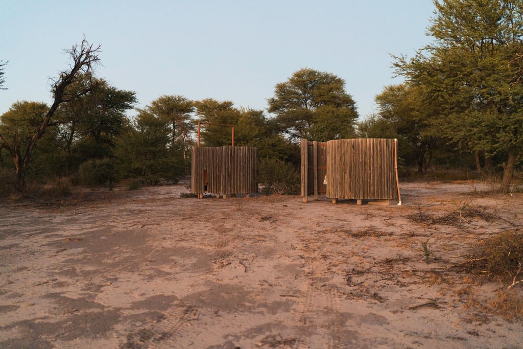 Zwei runde Holzkabinen auf sandigem Boden im Busch bei warmem Abendlicht.