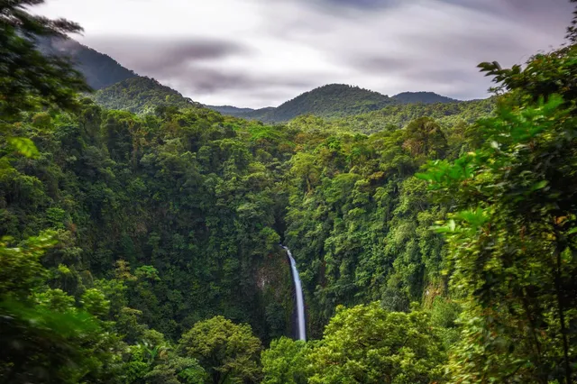 Blick in ein dicht bewaldetes Tal mit schmalem Wasserfall und bewaldeten Hügeln unter bedecktem Himmel.
