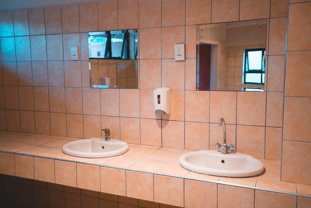Zwei Waschbecken mit Spiegeln und Seifenspender vor gefliester Wand, neutral beleuchtet.