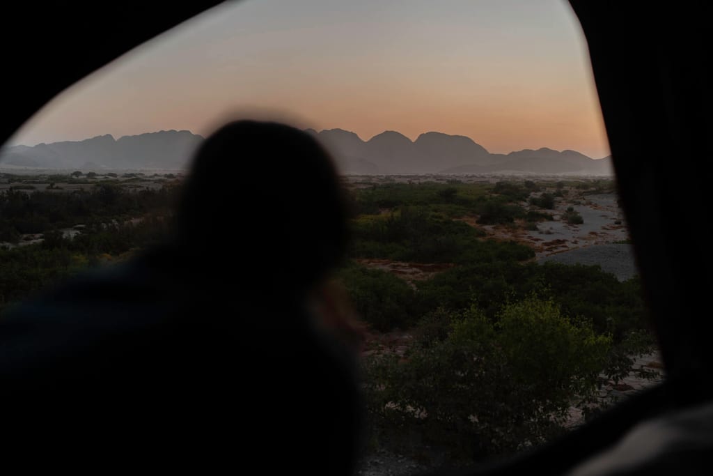 Silhouette im Fahrzeugfenster blickt bei Dämmerung auf Bergkette und trockene Flusslandschaft.