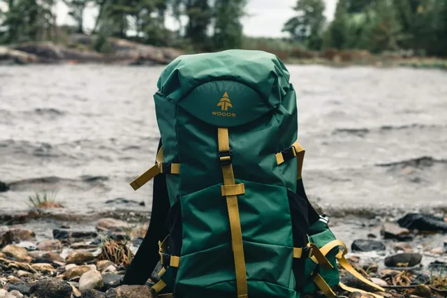 Grüner Wanderrucksack mit gelben Riemen auf steinigem Ufer vor ruhigem, bewölktem Gewässer.