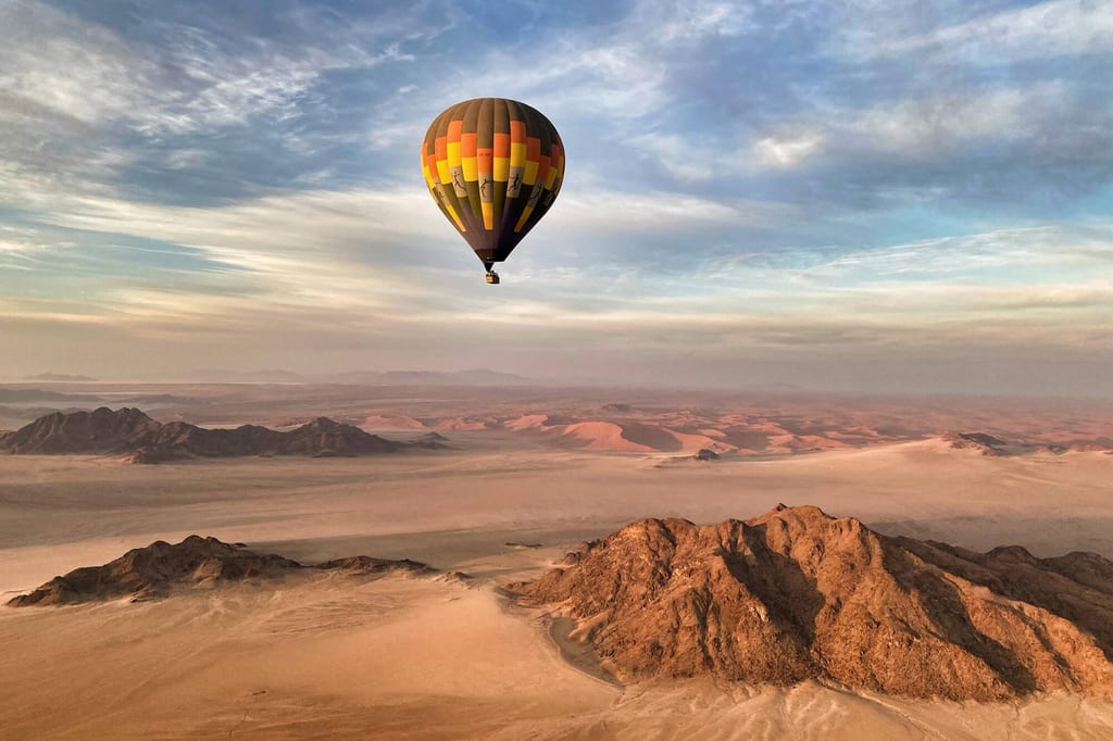 Heißluftballon über weiter Wüstenlandschaft mit Felsen und Dünen im warmen Morgenlicht.