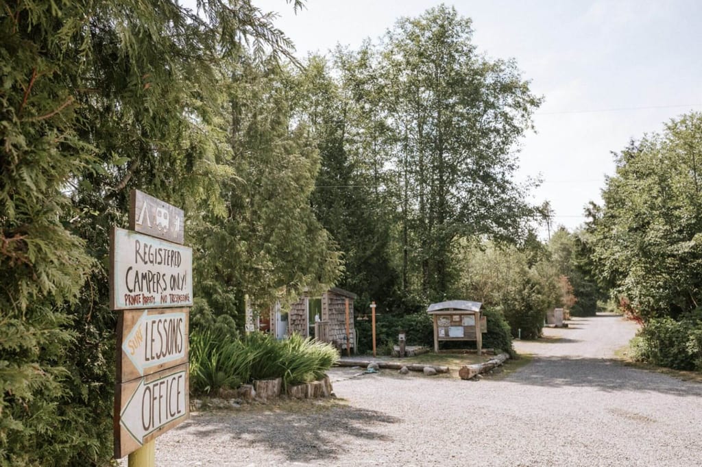 Eingang zu kleinem Campingplatz mit Schildern und geschotterter Zufahrt zwischen Bäumen