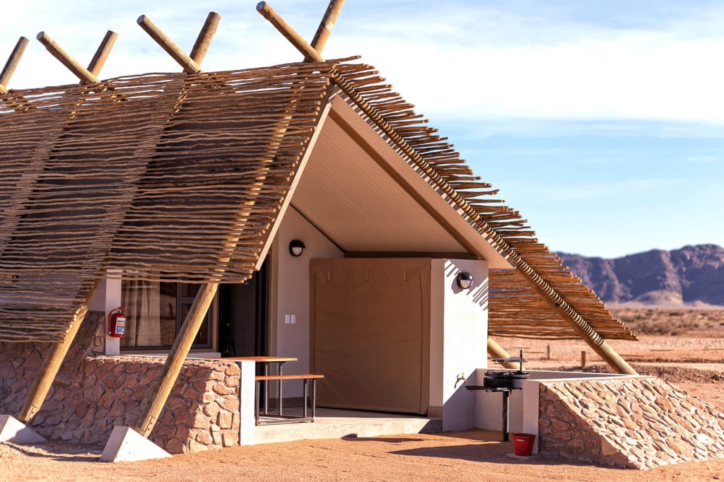 Kleine Lodge mit Holzsonnendach und Grill vor trockener Landschaft und Felsen