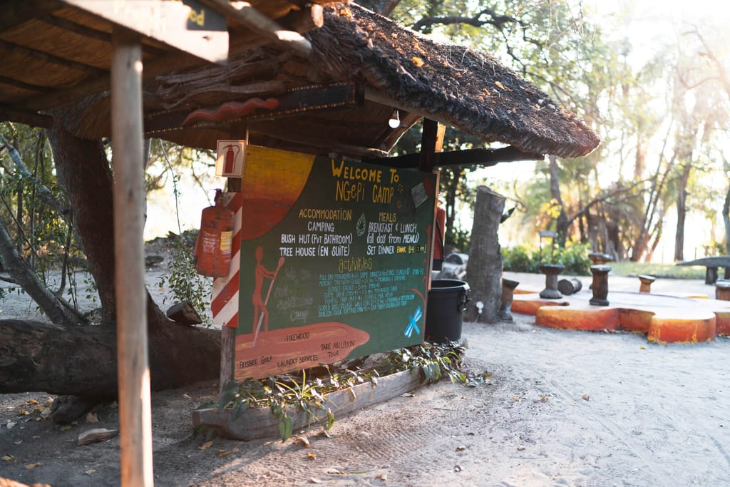 Handgemaltes Camp‑Schild unter Stroh­dach an sandigem Lagerplatz im warmen Abendlicht