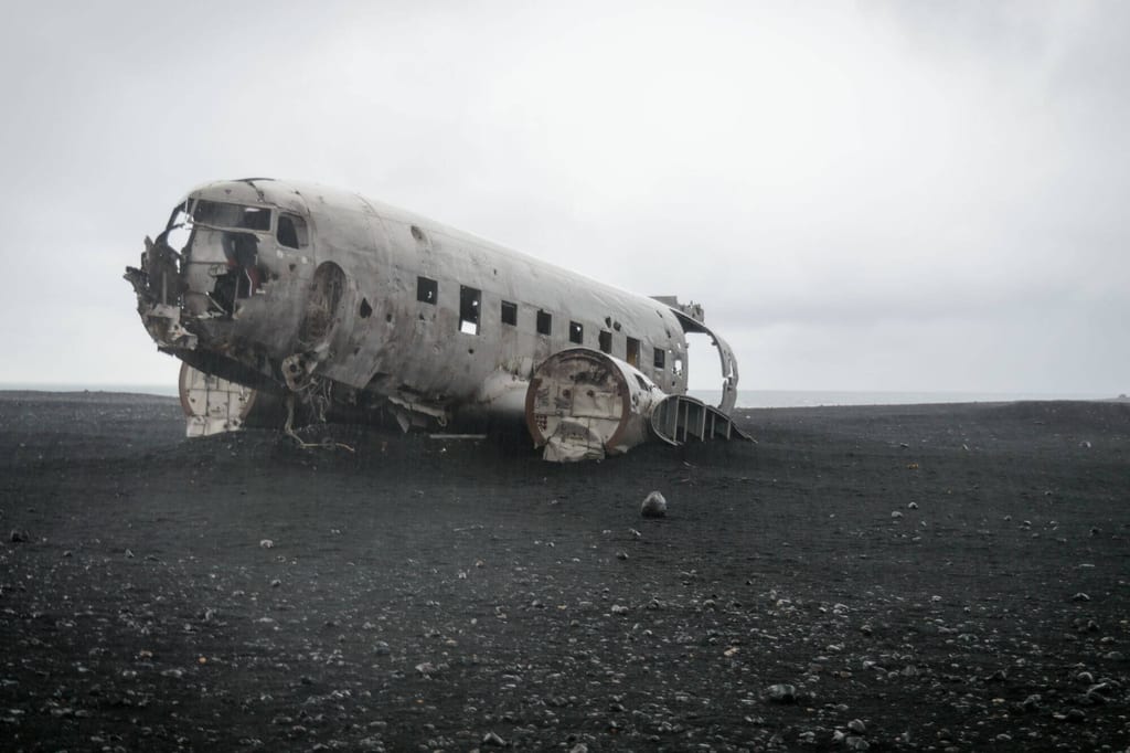Verlassenes Flugzeugwrack auf schwarzem Sand unter bedecktem Himmel, seitliche Frontalaufnahme
