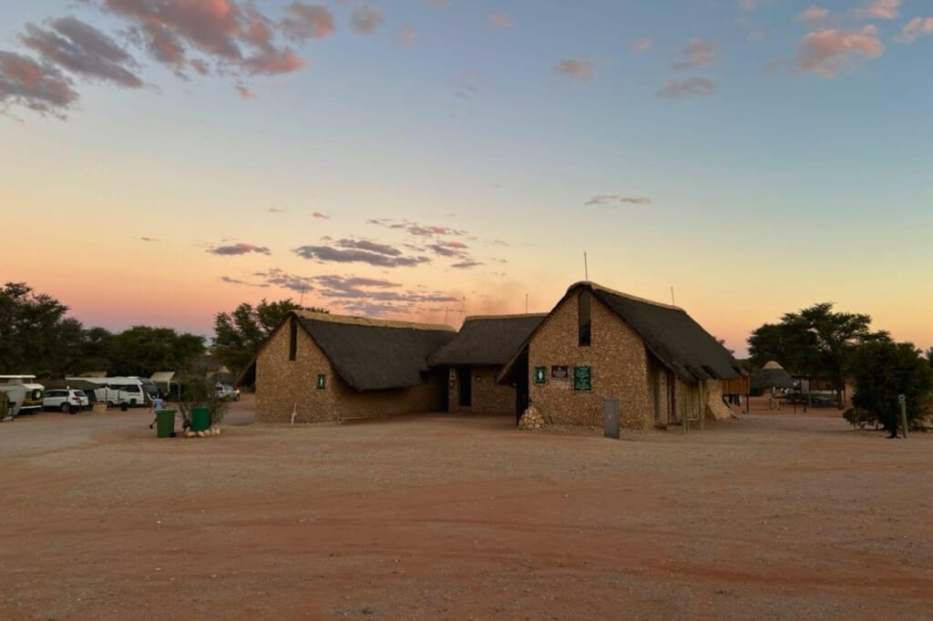 Strohgedeckte Gebäude und Camper auf staubiger Fläche bei pastelligem Sonnenuntergang