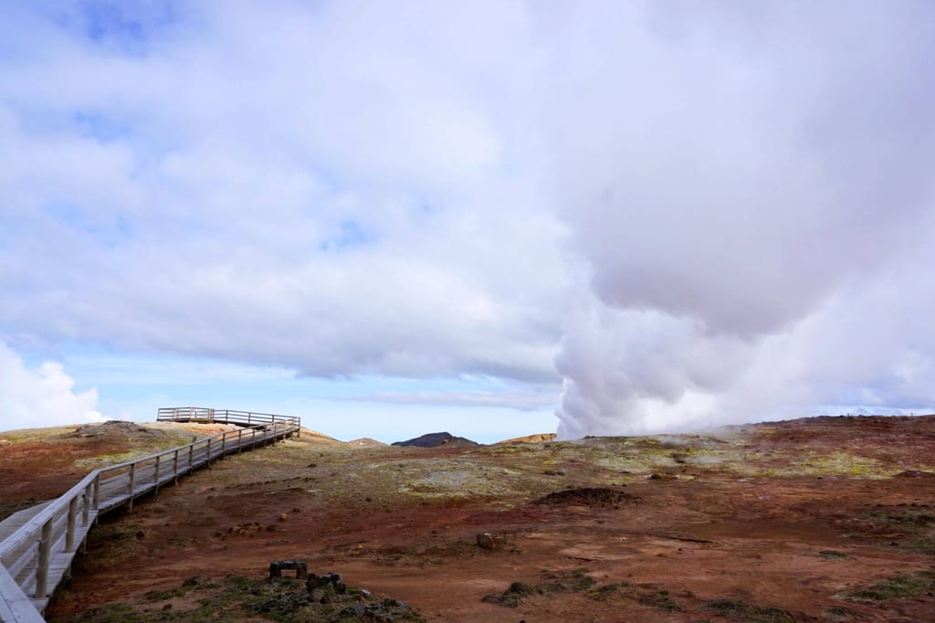 Hölzerner Steg führt durch karge Geothermielandschaft mit aufsteigender Dampfwolke bei bewölktem Himmel.