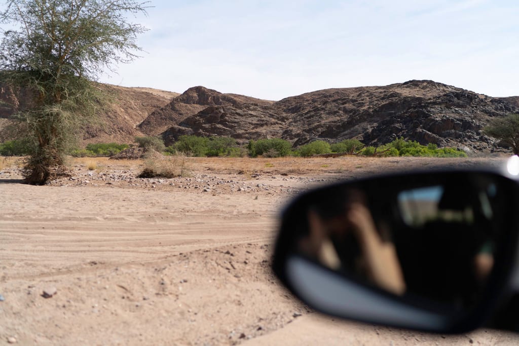 Blick aus einem Auto auf sandige Ebene mit Buschwerk und dunklen Felsen in Twyfelfontein, Namibia