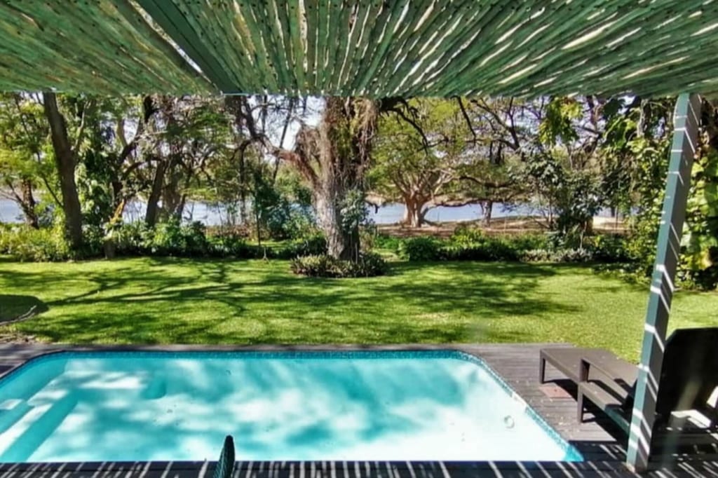 Blick von einer Pergola auf Pool, Rasen und Baumbestand am Ufer bei sonnigem Tageslicht