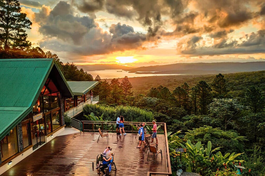 Berglodge-Terrasse mit Besuchern, Blick über Wald und See bei dramatischem Sonnenuntergang
