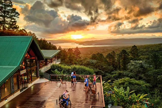 Berglodge-Terrasse mit Besuchern, Blick über Wald und See bei dramatischem Sonnenuntergang