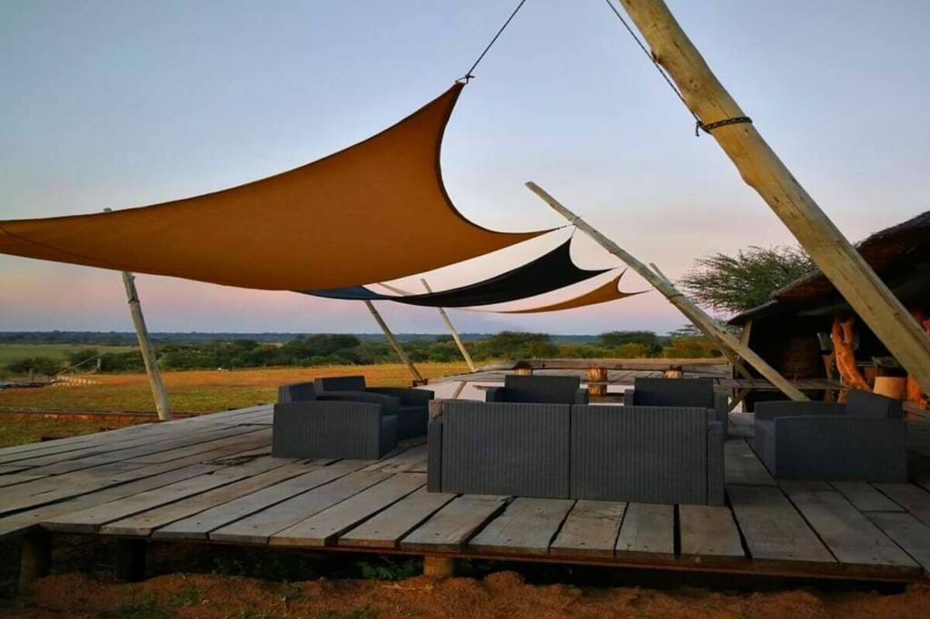 Holzdeck mit Lounge-Möbeln unter Sonnensegeln vor offener Savannenlandschaft im Abendlicht.