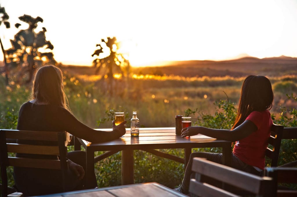 Zwei Personen sitzen bei Sonnenuntergang an einem Holztisch und schauen über eine offene Landschaft.