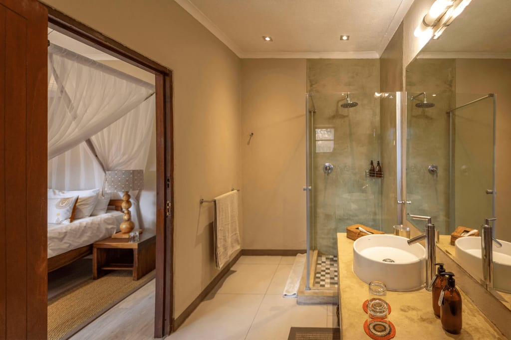 Warm beleuchtetes Badezimmer mit Glasdusche, rundem Waschbecken und Blick ins Schlafzimmer mit Himmelbett.