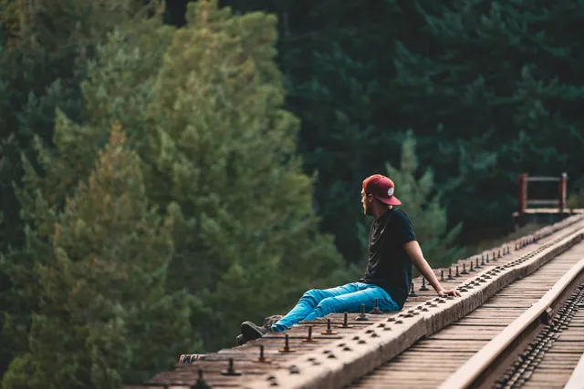 Person sitzt auf hölzerner Eisenbahnbrücke über dichtem Nadelwald, Blick seitlich nach rechts.