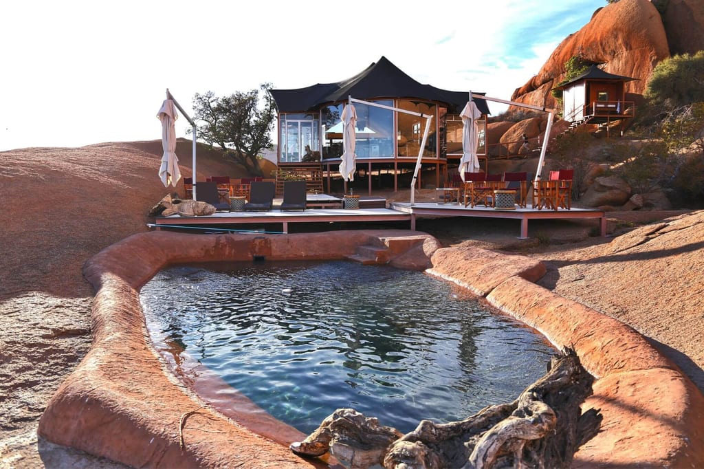 Zeltlodge mit Holzterrasse und Fels-Pool in roter Felsszenerie bei warmem Nachmittagslicht.