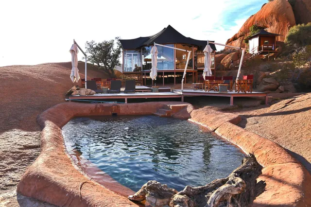 Zeltlodge mit Holzterrasse und Fels-Pool in roter Felsszenerie bei warmem Nachmittagslicht.
