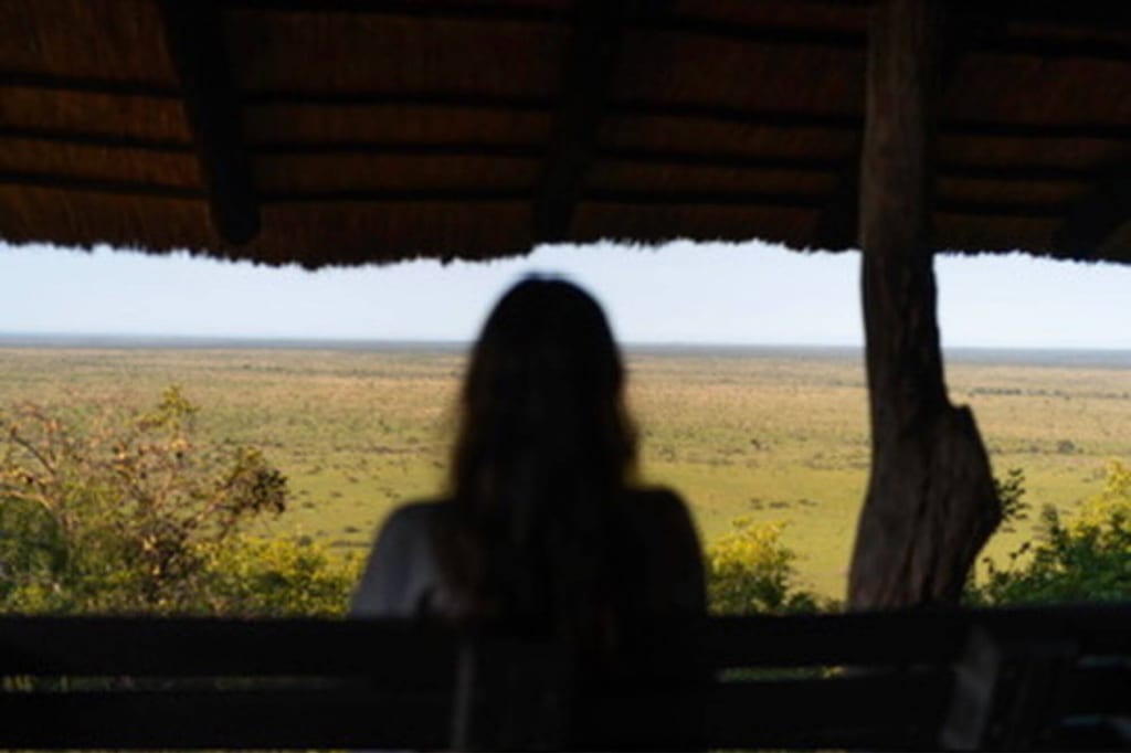 Person als Silhouette unter Strohdach blickt auf weite Graslandschaft am Tag