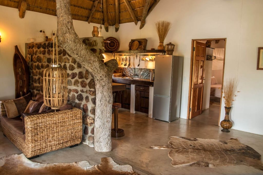 Rustikale Lodge innen mit Baumstammsäule, geflochtenem Sofa und kleiner Küche bei warmer Beleuchtung.
