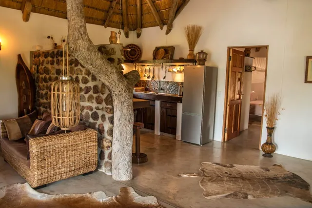 Rustikale Lodge innen mit Baumstammsäule, geflochtenem Sofa und kleiner Küche bei warmer Beleuchtung.