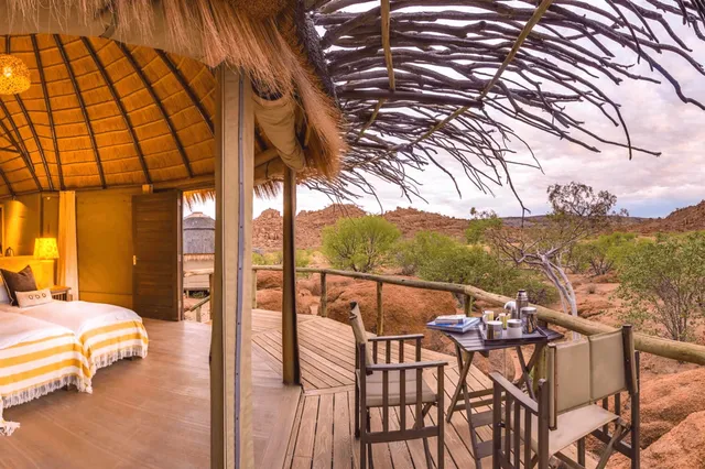 Offener strohgedeckter Bungalow mit Veranda und Tisch vor felsiger, buschiger Landschaft bei Dämmerung.