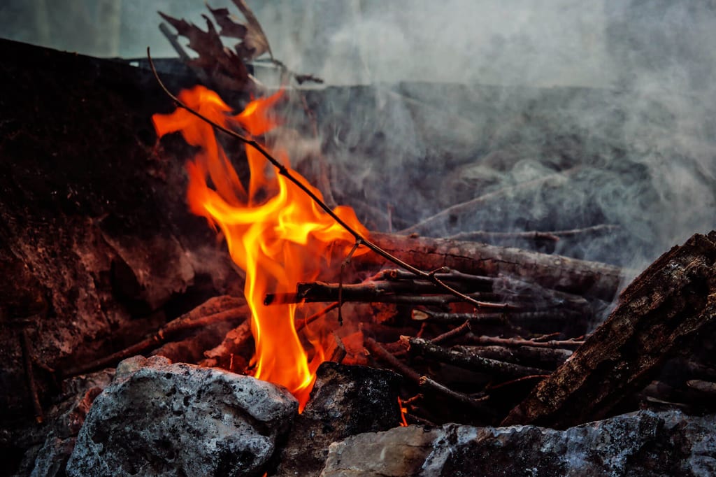 Nahaufnahme eines kleinen Lagerfeuers mit flackernden Flammen, Ästen und Rauch zwischen Steinen.