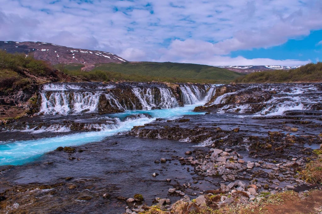 Mehrstufiger Wasserfall mit türkisblauem Fluss vor hügeliger, schneebedeckter Berglandschaft