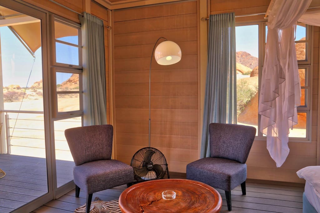 Gemütlicher Holzbungalow mit zwei Sesseln, rundem Tisch und Blick auf felsige Wüste durch große Fenster.