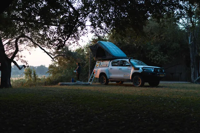 Geländewagen mit aufgeklapptem Dachzelt und Person am Waldrand in der Abenddämmerung.