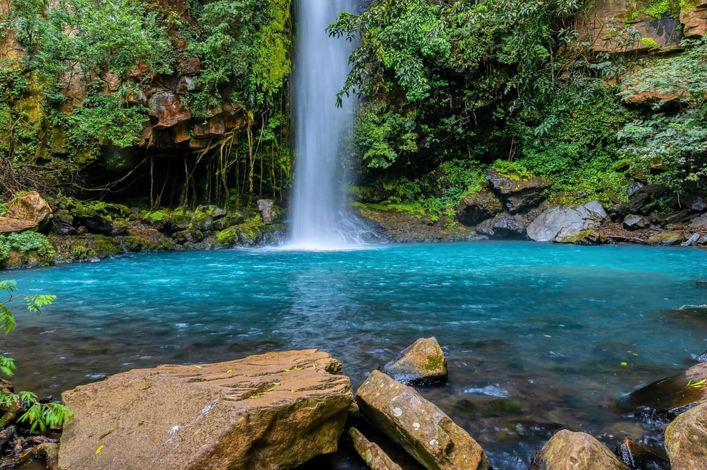 Wasserfall fällt in türkisblaues Becken, umgeben von moosigen Felsen und dichter grüner Vegetation.