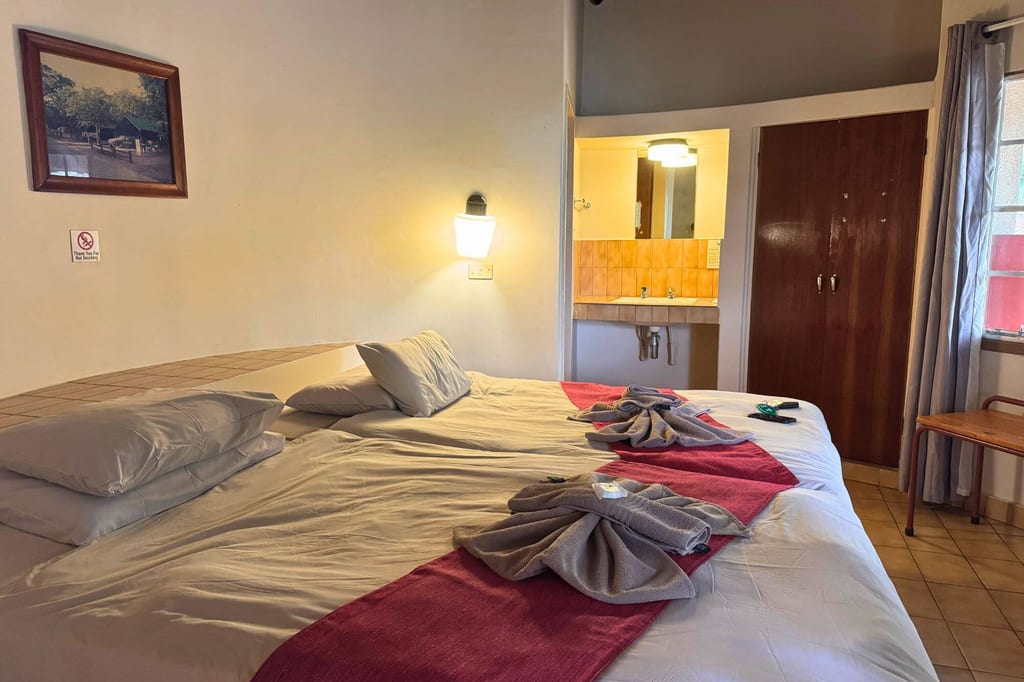 Einfaches Hotelzimmer mit großem Bett, gefalteten Handtüchern und offenem Waschbereich, warme Beleuchtung.