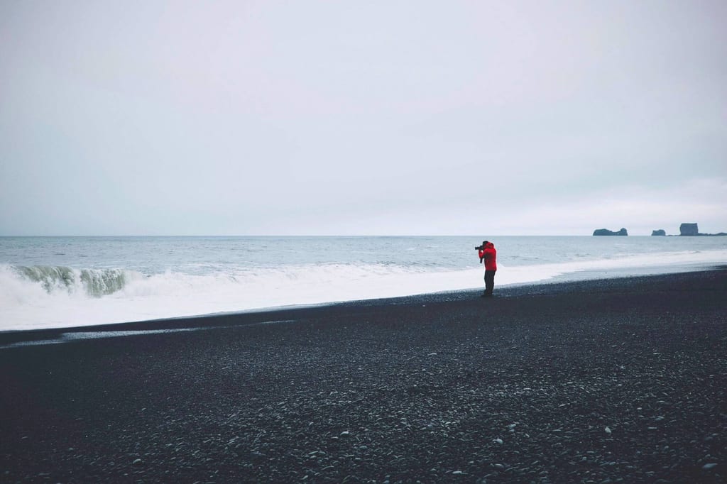 Person in roter Jacke fotografiert das Meer an einem schwarzen Kiesstrand bei bedecktem Himmel.