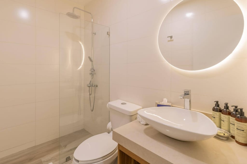 Modernes Badezimmer mit Waschbecken, WC, gläserner Dusche und rundem hinterleuchtetem Spiegel.