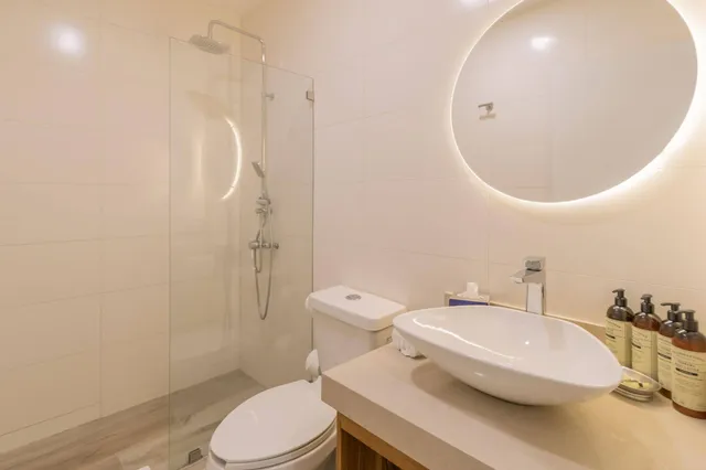 Modernes Badezimmer mit Waschbecken, WC, gläserner Dusche und rundem hinterleuchtetem Spiegel.