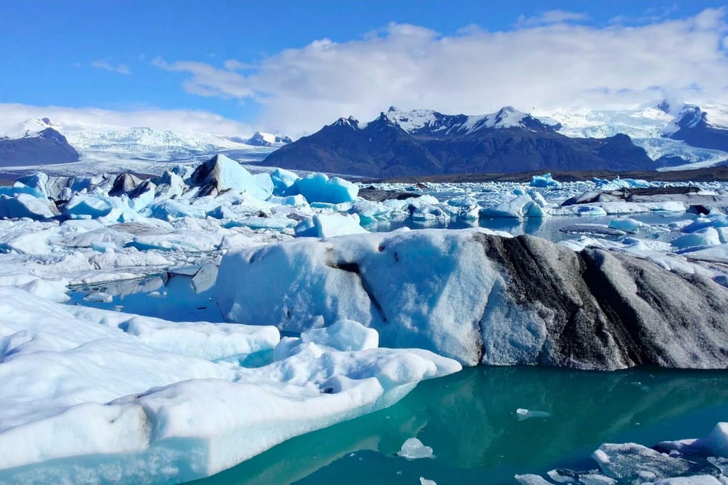 Eislagune mit treibenden Eisbergen, türkisem Wasser und Bergen im Hintergrund bei klarem Sonnenschein
