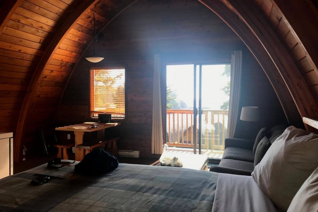 Innenansicht einer Holz‑A‑Frame‑Hütte mit Bett, Sofa und Balkonblick bei warmem Tageslicht.