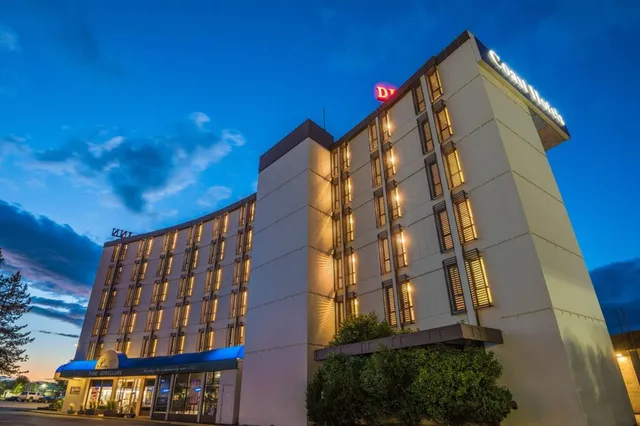 Mehrstöckiges Hotel bei Dämmerung mit warm beleuchteten Fenstern und Sträuchern im Vordergrund.