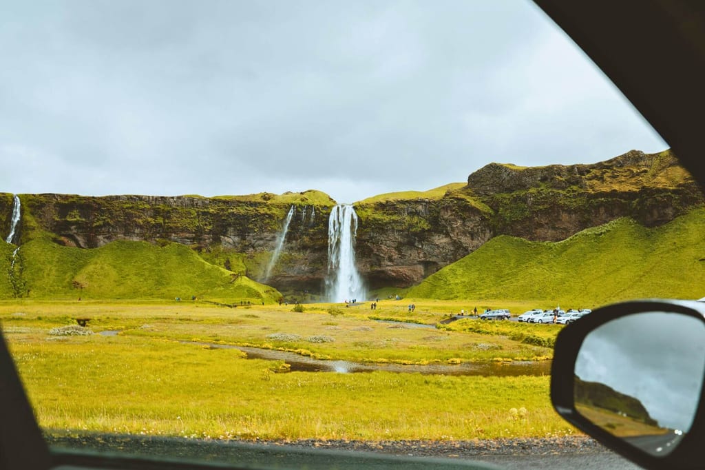 Blick aus dem Auto auf einen hohen Wasserfall in grüner Hügellandschaft bei bewölktem Tageslicht.