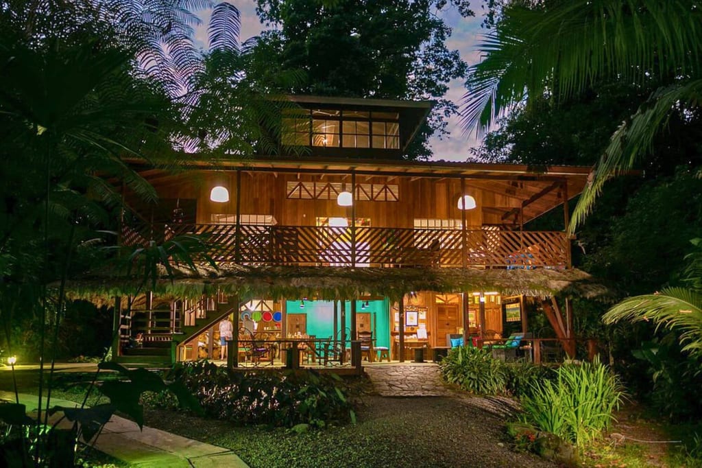 Beleuchtete Holzlodge mit großer Veranda in dichter tropischer Vegetation bei Abenddämmerung.