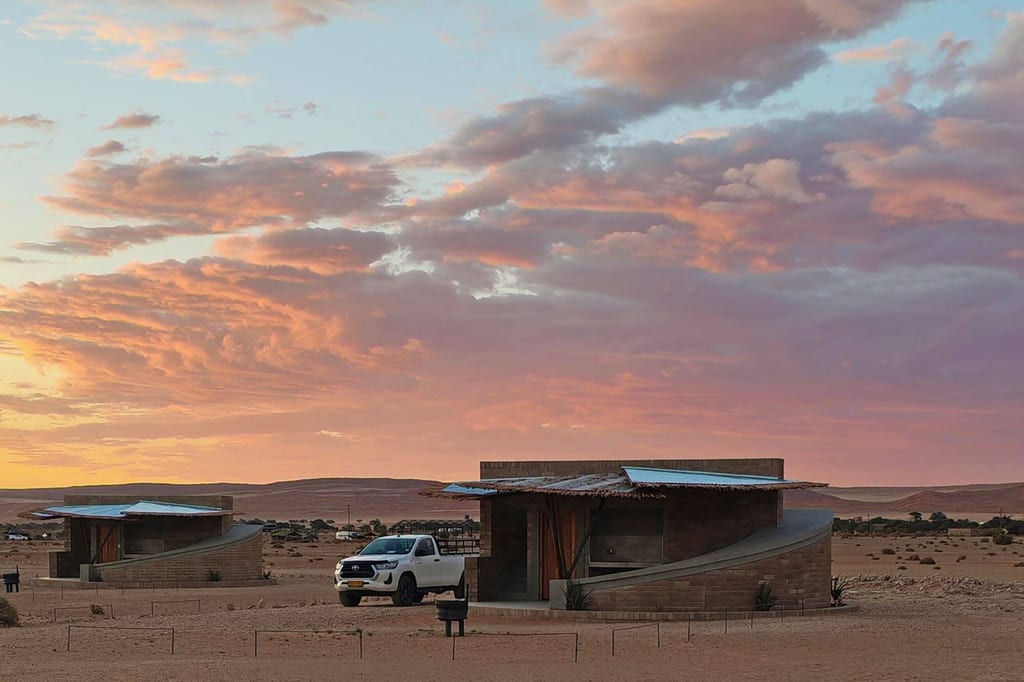 Zwei Wüstenbungalows mit weißem Pickup vor rosa-violettem Sonnenuntergangshimmel.
